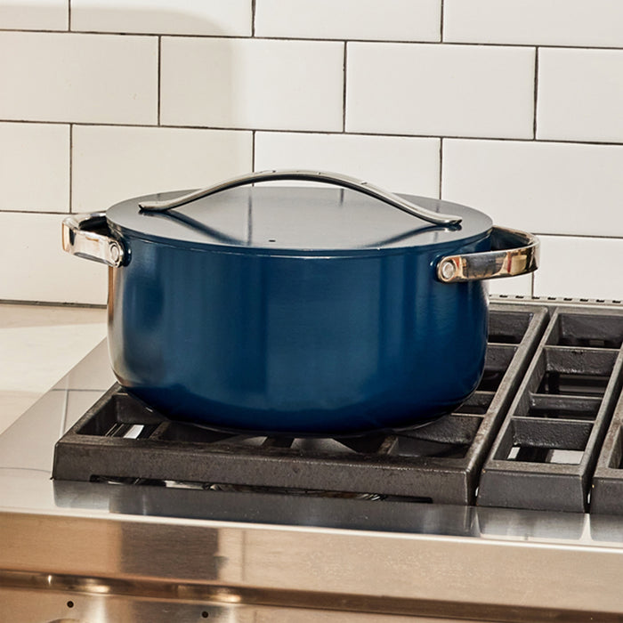 A Caraway Ceramic Dutch Oven, 6.5 qt, with a blue lid and shiny metal handles sits on a gas stovetop atop a stainless steel surface, framed by white subway tiles.