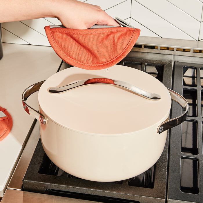 A hand uses an orange pot holder to lift the lid of a 6.5 qt ceramic-coated, non-toxic Caraway Dutch Oven on the stove against a modern kitchen with a white geometric tiled backsplash.