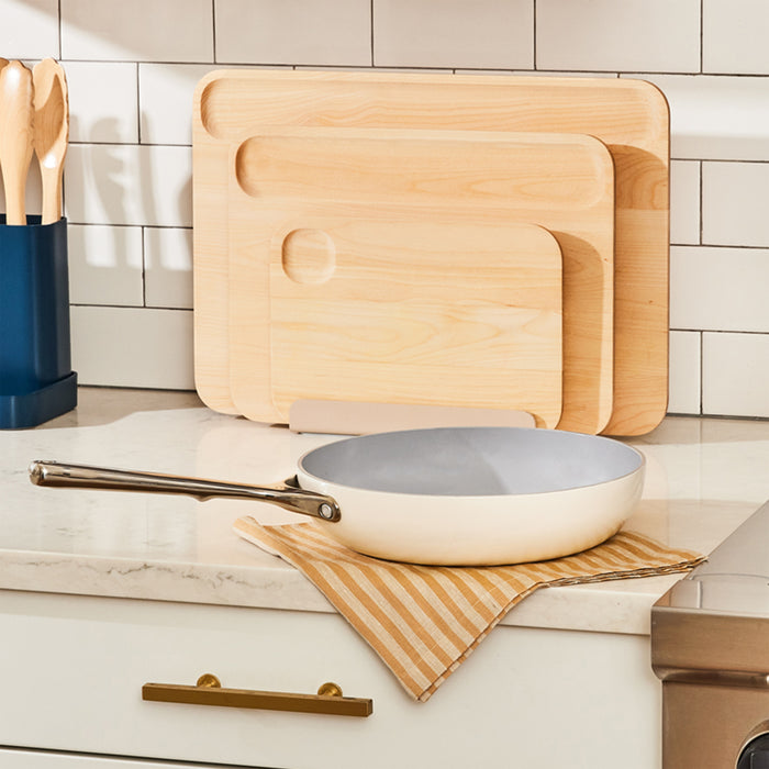 A kitchen scene features three wooden cutting boards against a white tiled backsplash. A Caraway 10.5 Ceramic Fry Pan with a silver handle sits on a striped cloth atop a marble countertop, while wooden utensils are stored in a blue holder.