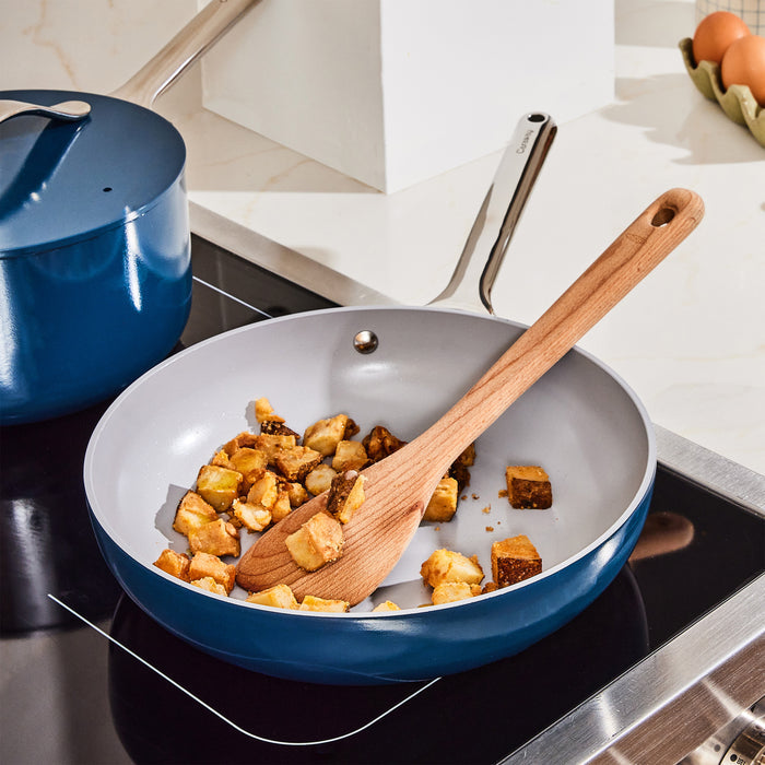 The Caraway 10.5 Ceramic Fry Pan, featuring a naturally non-stick surface and wooden handle, stirs food cubes on an induction stove. In the background, a blue pot with a lid is beside a carton of eggs on the white countertop.