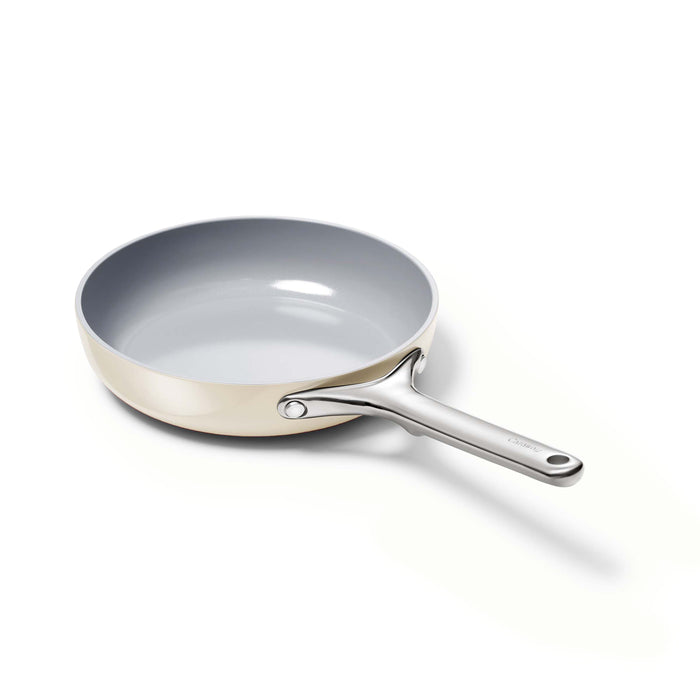 The Caraway 8 Ceramic Fry Pan features a non-stick surface with a grey interior and light-colored exterior. It has a sleek metal handle secured by two rivets and is presented tilted against a white background.