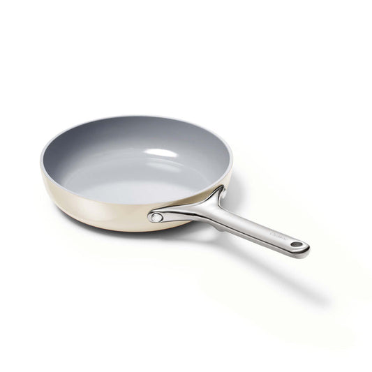 The Caraway 8 Ceramic Fry Pan features a non-stick surface with a grey interior and light-colored exterior. It has a sleek metal handle secured by two rivets and is presented tilted against a white background.