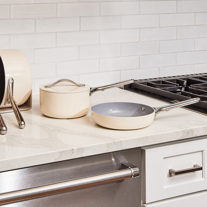 A modern kitchen countertop features a cream-colored Caraway Ceramic Fry Pan, 8 and a saucepan with lids. Nearby, cutting boards lean against white subway tile backsplash. A stainless steel gas stove is partially visible.