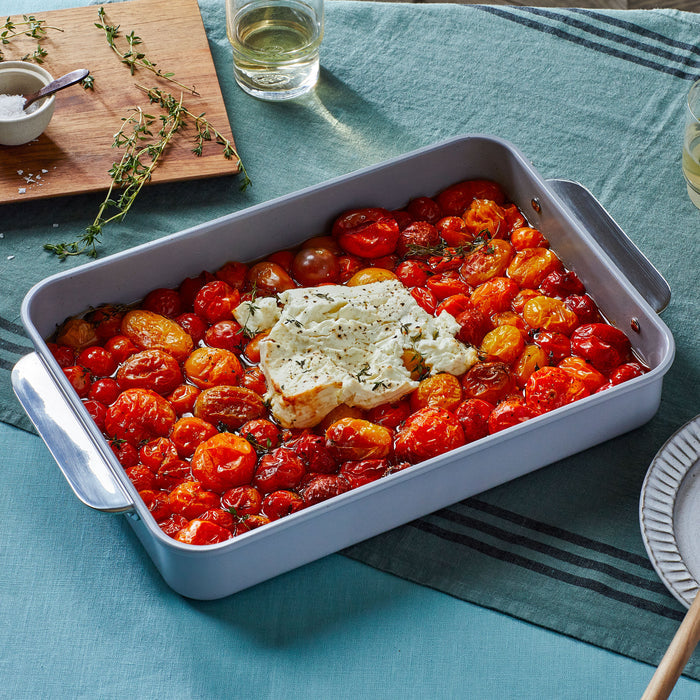 A Ceramic Rectangle Baking Pan by Caraway, filled with colorful roasted cherry tomatoes and cheese, rests on a teal cloth. Nearby, a non-stick pan with a ceramic coating sits next to a wooden board topped with herbs, white wine glasses, and plates.