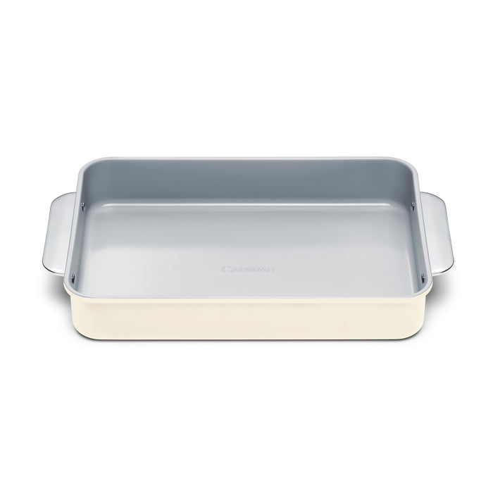 The Caraway Ceramic Rectangle Baking Pan, featuring a non-toxic ceramic coating, is beige with a metallic gray interior and rounded edges. It has wide handles for easy use and is showcased on a plain white background.
