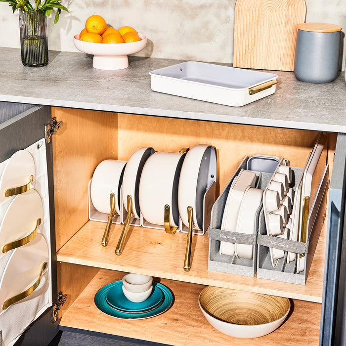 The open kitchen cabinet displays neatly organized white pots, pans, and Caraways Ceramic Rectangle Baking Pan with gold handles alongside a non-stick pan. On the countertop, a fruit bowl with oranges and small wooden cutting boards are lit up in bright light, accentuating the modern design.