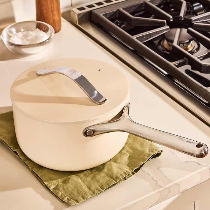 A cream-colored Caraway Ceramic Sauce Pan, 3 qt with a silver handle rests on a green cloth atop a marble countertop. The lid is closed, and a gas stove is visible in the background. Nearby, a small bowl of salt awaits use.