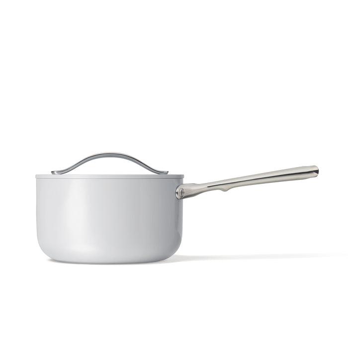 The Caraway Ceramic Sauce Pan, 3 qt, is a stylish white saucepan with a lid and long silver handle, featuring a ceramic-coated interior. It rests elegantly against a plain white background, blending aesthetics with functionality.