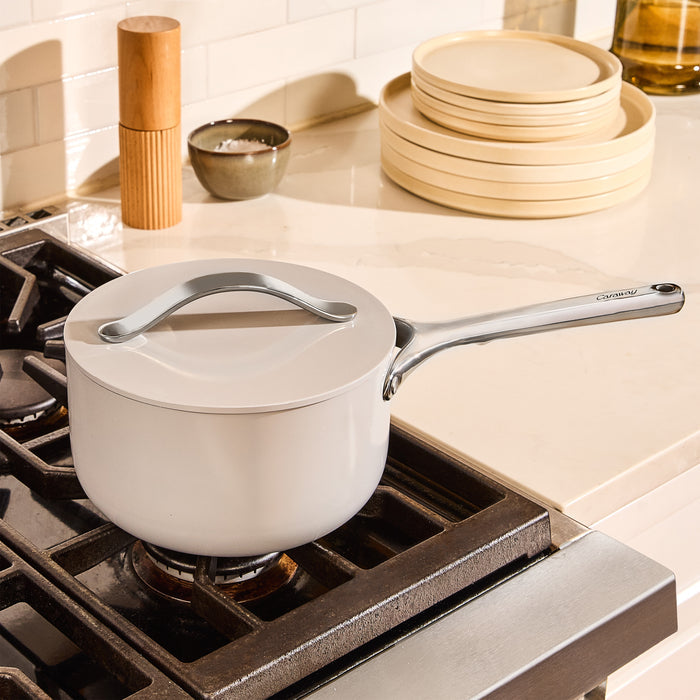 A Caraway Ceramic Sauce Pan, 3 qt with a lid rests on a stove burner. In the background, a stack of beige plates, a salt container, and a wooden pepper mill adorn the light-colored countertop.
