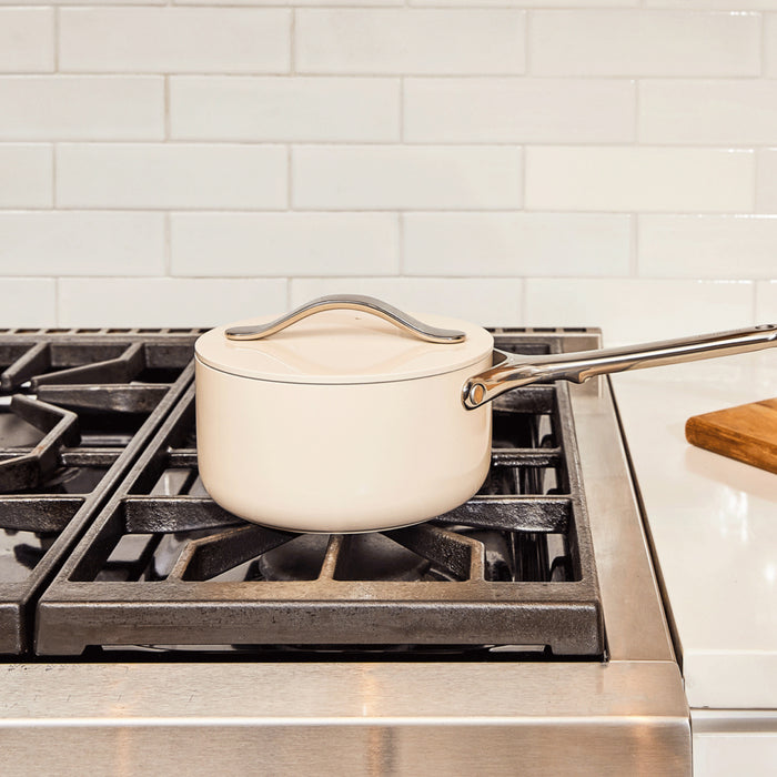 A Caraway Ceramic Sauce Pan with Lid, 1.75 qt, rests on a gas stove, praised for healthy cooking benefits. The black grates contrast with the white subway tile backsplash, while a wooden cutting board peeks from the right, adding warmth to this culinary setting.