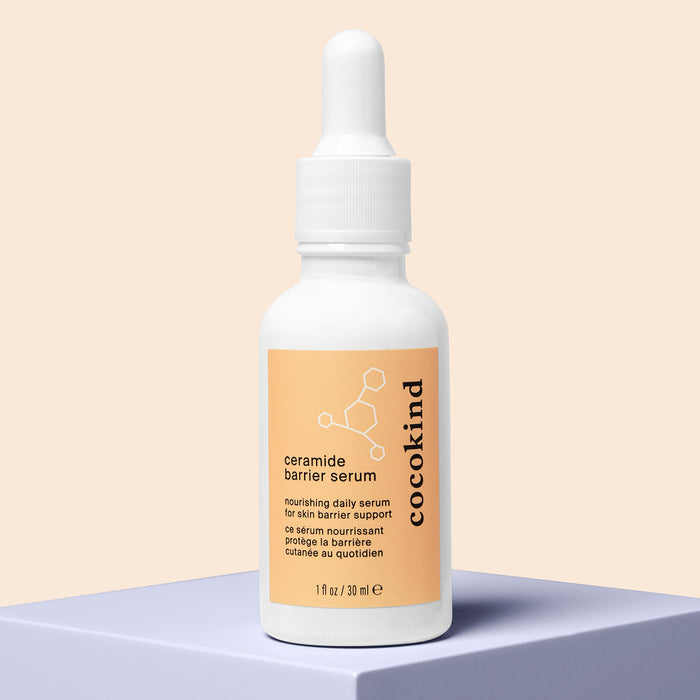 A Cocokind Ceramide Barrier Serum bottle, featuring a white design with an orange label and molecular graphics, sits on a blue platform, highlighting its role in nourishing and supporting the skins natural barrier.