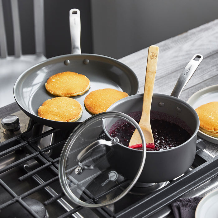 Three pancakes sizzle in a PFAS-Free frying pan while nearby, blueberry sauce simmers in a GreenPan Chatham Ceramic Non-Stick saucepan. A wooden spoon stirs the sauce as a glass lid tops this Thermolon nonstick marvel on the modern kitchen stove.
