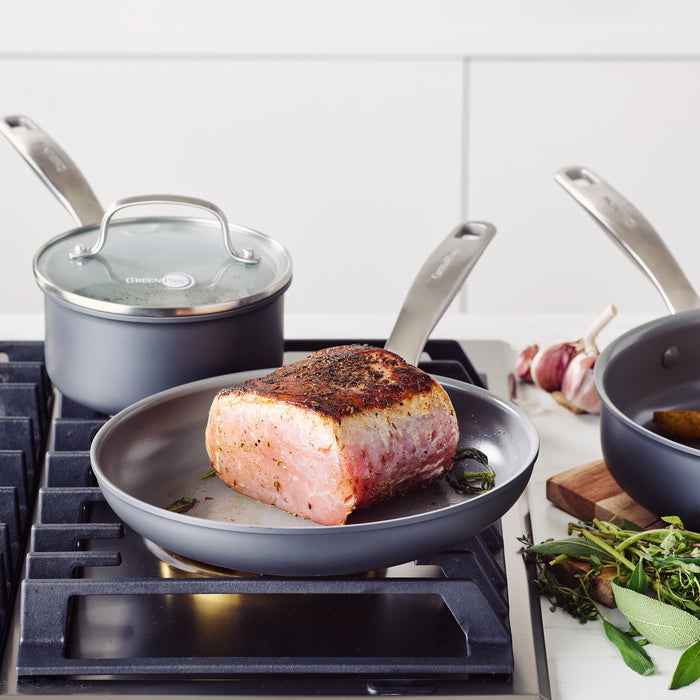Meat sizzles in a frying pan beside GreenPans Chatham Ceramic, Non-Stick Cookware Set with a glass-lid saucepan. Fresh herbs and garlic bulbs promise aromatic bliss—all while ensuring peace of mind with PFAS-Free cookware.