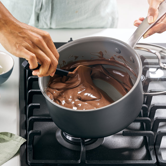 A person stirs melted chocolate on a gas stove using their GreenPan Chatham Ceramic Non-Stick Cookware, featuring its renowned Thermolon ceramic nonstick surface. Nearby, a green cloth and small bowl accent the scene, as they craft with PFAS-free cookware in a light-colored shirt.