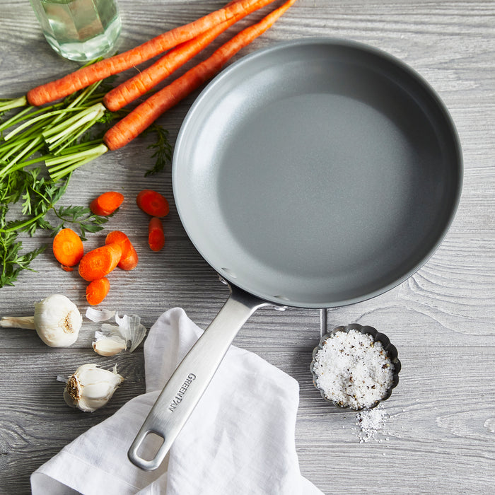 A Chatham Nonstick Ceramic Fry Pan by GreenPan sits on wood, surrounded by whole and sliced carrots, garlic cloves, coarse salt in a small dish, and a white cloth. A glass jar appears at the top left corner.