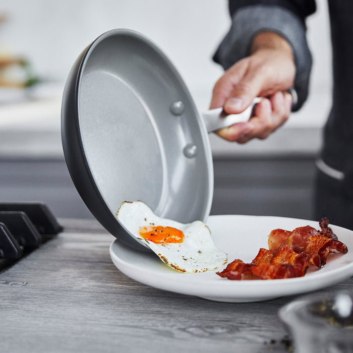 In a kitchen, someone slides a sunny-side-up egg from the GreenPan Chatham Nonstick, Ceramic Fry Pan, 8 onto a white plate with bacon. The PFAS-Free cookware gleams under the gentle stove light.