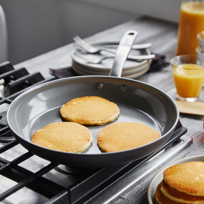 Three golden pancakes sizzle in GreenPans Chatham Nonstick, Ceramic Fry Pan, 8, featuring a PFAS-free ceramic nonstick coating. In the background, stacked plates, silverware, and a glass of fresh orange juice create an inviting scene on the wooden table.