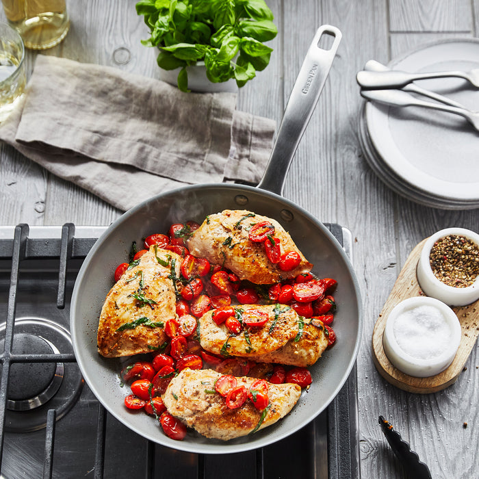 On the stovetop, a GreenPan Chatham Nonstick Ceramic Fry Pan holds four chicken breasts with tomatoes and herbs. Nearby are a salt and pepper set, stacked plates with cutlery, a glass of white wine, a napkin, and basil plant. Its PFAS-free design ensures healthy cooking.