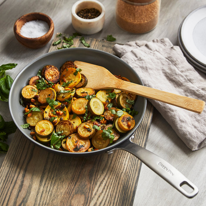Sautéed sliced zucchini with herbs in a GreenPan Chatham 8 Nonstick Ceramic Fry Pan, PFAS-Free, rests on a wooden table. A wooden spatula lies in the pan. Nearby are bowls of salt, pepper, brown sugar, and a stack of white plates with a linen napkin.