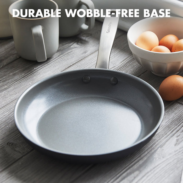 The GreenPan Chatham Open Frypan, 12 in, PFAS free with a hard anodized body and metal handle, sits on a wooden table next to a white bowl of eggs and mugs. Text above reads: DURABLE WOBBLE-FREE BASE.