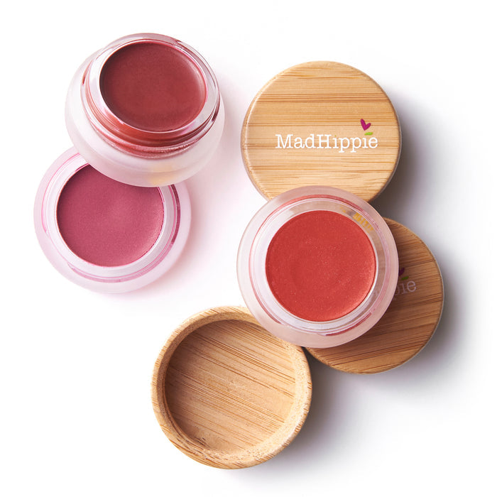 Three pots of Mad Hippie Cheek & Lip Tint with bamboo lids are displayed on a white background. One pot is open to reveal another shade, and the balms serve as both cheek and lip tint in pink and red tones.
