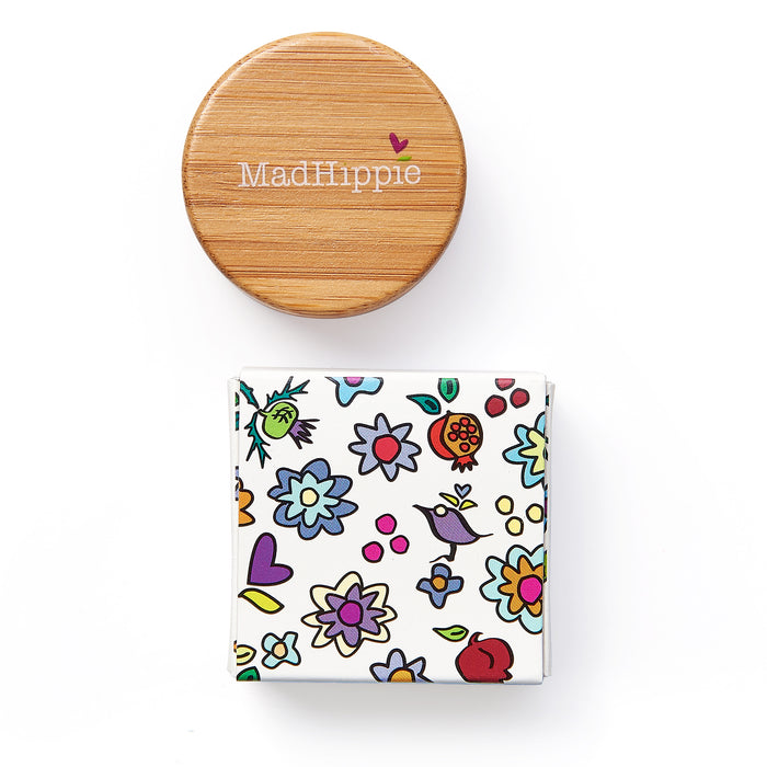 A round wooden lid labeled Mad Hippie lies beside a vibrant box with floral illustrations. This playful design showcases the Cheek & Lip Tint, emphasizing a natural theme ideal for eco-conscious users exploring Mad Hippies recycling program.