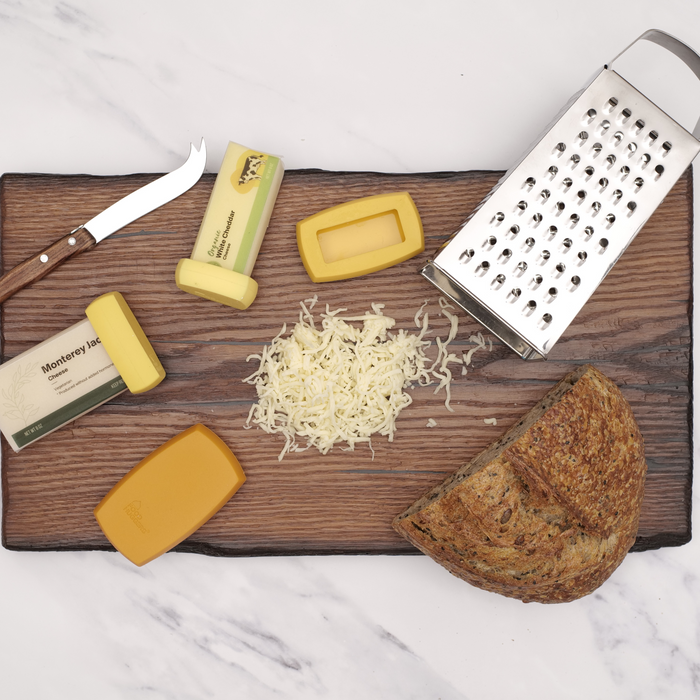 A large cutting board showcasing Cheese Huggers, hard cheese blocks, shredded cheese, a knife, grater, and multigrain bread on marble suggests a space-saving setup for preparing a cheesy meal. Branding by Food Huggers ties it all together.