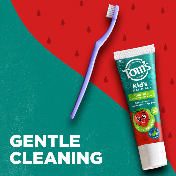 A tube of Tom's of Maine Kids' Natural toothpaste in Silly Strawberry flavor lies on a green and red background with a blue toothbrush beside it. White text reads "Gentle Cleaning." Raindrop-like designs are scattered on the red section.