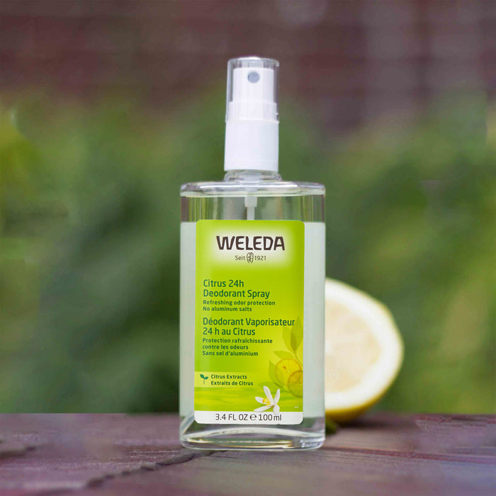 A 100ml bottle of Weledas Citrus 24h Deodorant Spray sits on wood, backed by a fresh lemon half. The aluminum-free green label, decorated with citrus designs, is highlighted by the blurred background emphasizing its citrus scent.