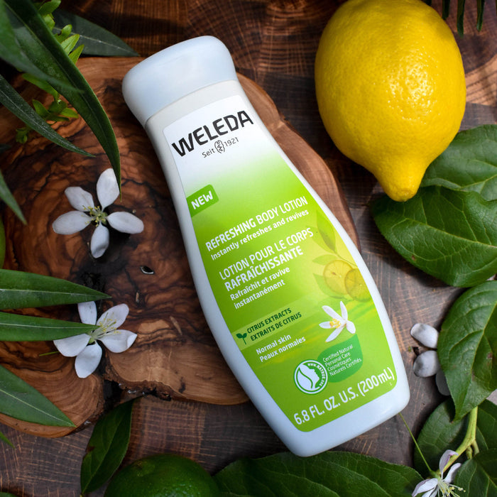 A bottle of Weleda Citrus Refreshing Body Lotion sits among green leaves, a lemon, small white flowers, and wooden accents. The green and white label showcases citrus extracts and natural origin ingredients like soothing aloe vera for a revitalizing experience.
