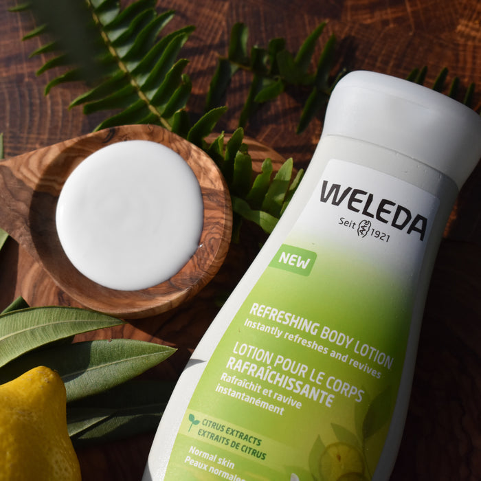 A bottle of Weledas Citrus Refreshing Body Lotion sits on a wooden surface, surrounded by green leaves and a lemon. A small bowl holds the hydrating lotion enriched with aloe vera, highlighting natural freshness and botanical elements in the bright scene.