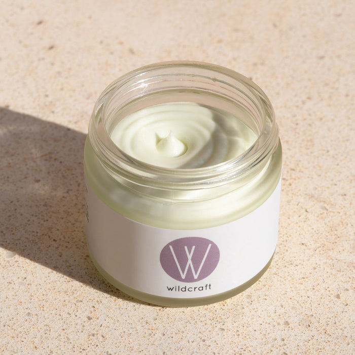 A glass jar of Clarify Face Cream by Wildcraft, featuring a lavender W logo, sits on a textured beige surface. The light green cream is ideal for sensitive skin and has a swirled peak in the center.