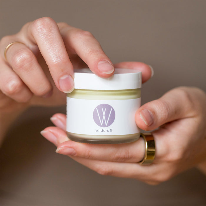 Hands gently cradle a jar of Wildcraft Clarify Face Cream with a white lid and purple logo. The person is carefully opening the jar, ideal for sensitive or dry skin, against a neutral background.