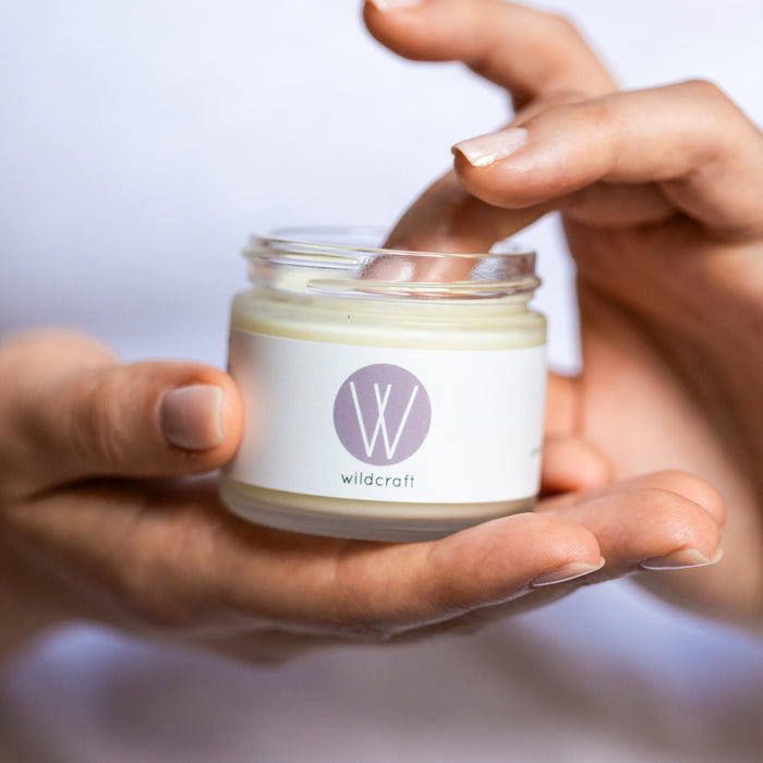 A person with manicured nails dips a finger into a small Clarify Face Cream jar from Wildcraft, featuring a stylized W logo. Perfect for sensitive skin, the focus is on the hands and the jar against a simple background.