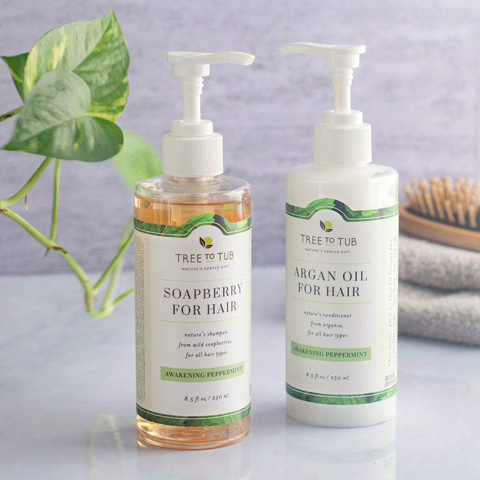 Two Tree To Tub hair care bottles, the Clarifying Shampoo for Oily Hair & Sensitive Scalp and “Argan Oil for Hair” conditioner, are elegantly displayed on a light surface. A plant, towel, and wooden brush add charm in the background—ideal for a sensitive scalp routine.