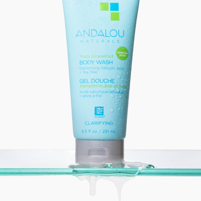 The image features a tube of Andalou Naturals Clarifying Yuzu Grapefruit Body Wash on a wet surface. The light blue tube, showcasing a green and white label, displays this clean skincare product as detoxifying gel drips from it onto the surface.