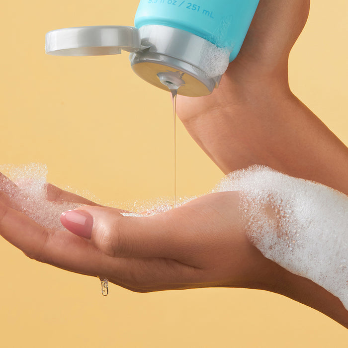 A person pours Andalou Naturals Clarifying Yuzu Grapefruit Body Wash from a teal bottle onto their hand, creating white foam. The soft yellow background evokes clean skincare and serenity.