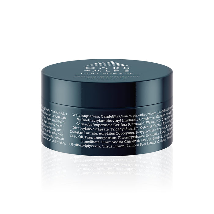 A dark teal jar of Oars + Alps Clay Pomade with a white mountain logo on the lid and detailed ingredient list on the side offers medium matte hold for stronger, healthier hair. The reflected jar sits on a white background.