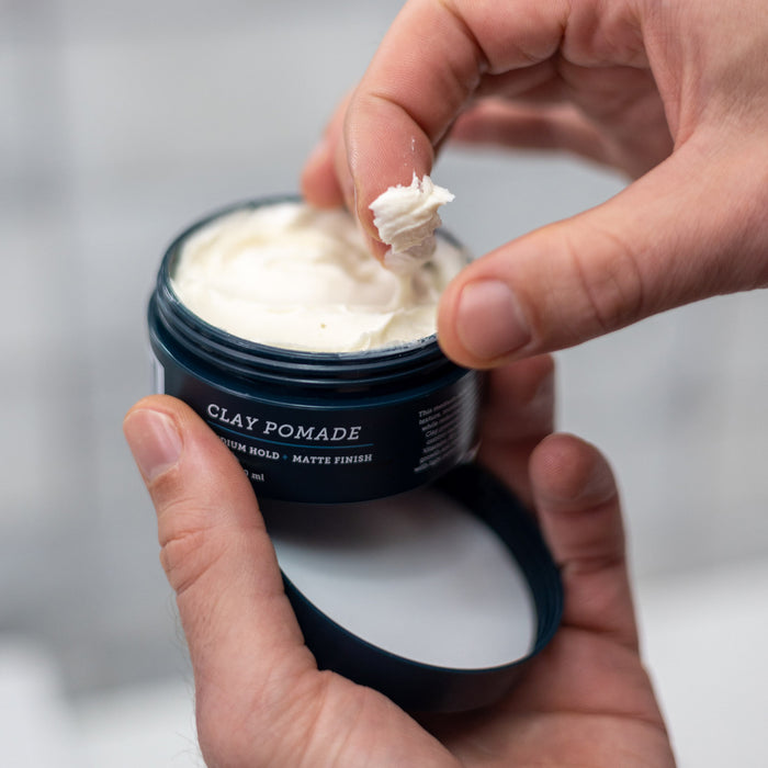 A person scoops Oars + Alps Clay Pomade from an open black container, preparing to use their fingers for a frizz-free style and medium matte hold.