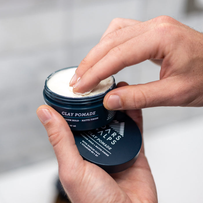 Someone scoops Oars + Alps Clay Pomade from a blue jar labeled Clay Pomade with their fingers, holding the container in the other hand. The blurred background highlights this medium matte hold formula for frizz-free style.
