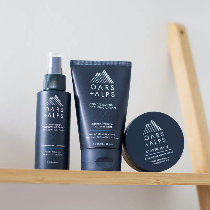 Oars + Alps Clay Pomade, designed for a frizz-free style with medium matte hold, is neatly arranged on a light wooden shelf against a plain background.