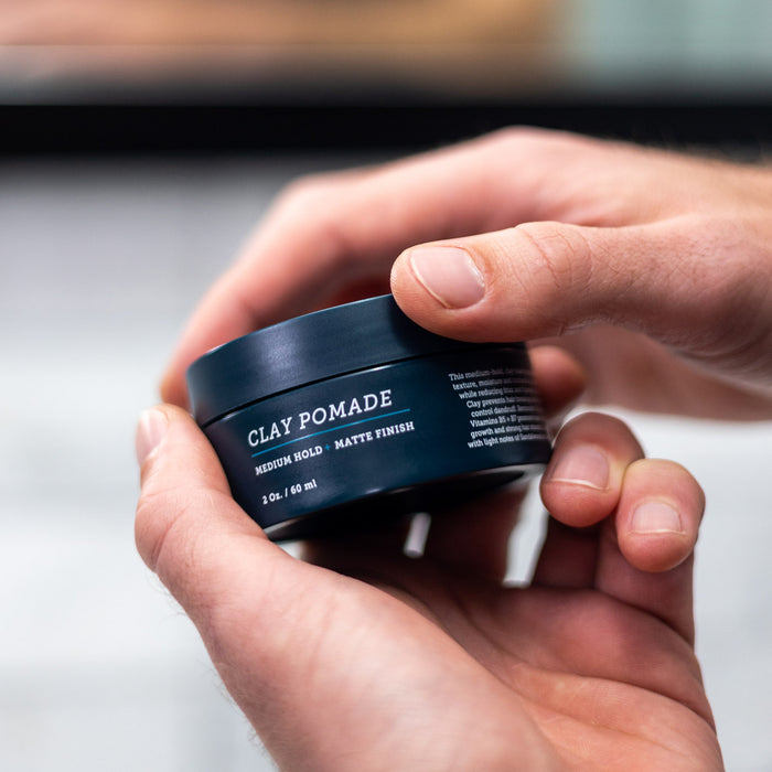 A person holds an Oars + Alps Clay Pomade container labeled Medium Hold · Matte Finish, promising frizz-free style, with a blurred background.