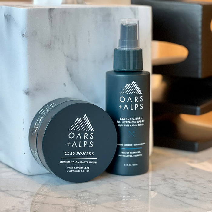 Oars + Alps Clay Pomade, with its sleek dark blue packaging and white logo, sits on a marble surface beside a wooden tray, offering a frizz-free style and medium matte hold.