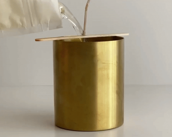 A hand pours wax from the Siblings DIY Candle Kit into a golden holder, securing a wooden wick with a popsicle stick. The setup of clean ingredients is on a pristine white surface.