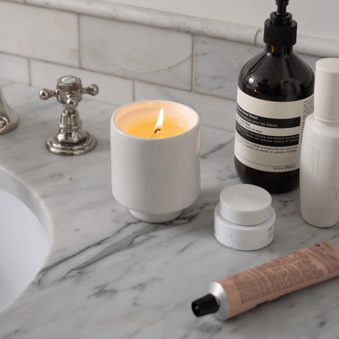 A lit DIY Candle Kit by Siblings in a white holder sits on a marble bathroom counter, surrounded by a faucet, hand soap dispenser, cream jars, and lotion. Made with clean ingredients and packaged in compostable material, the scene is neutral and minimalist.