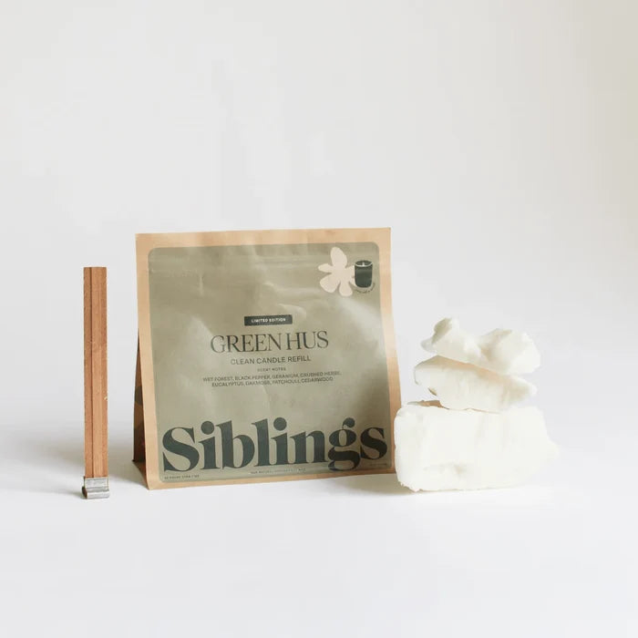 A Siblings DIY Candle Kit is shown: a green refill pouch stands upright beside a wooden wick and three pieces of white wax, all against a plain white background—ideal for eco-friendly candle making with compostable packaging.