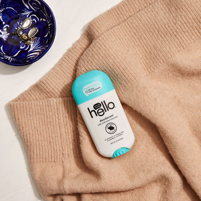 A stick of hello Clean + Fresh Deodorant with Activated Charcoal and shea butter rests on a beige sweater. Next to it, a blue and white dish with jewelry sits on a white surface, showcasing the deodorants all-natural, recyclable packaging.