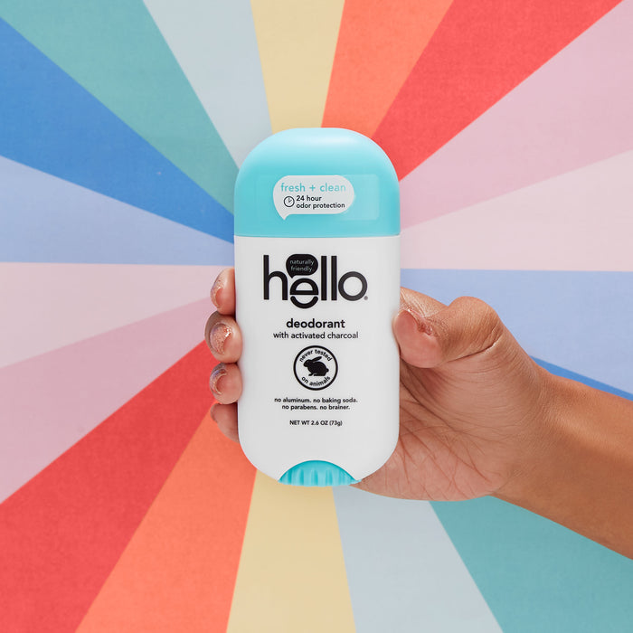 A hand holds hellos Clean + Fresh Deodorant with Activated Charcoal, showcasing an all-natural choice. The white and turquoise stick stands out against a backdrop of pastel rainbow rays, emphasizing the recyclable packaging.