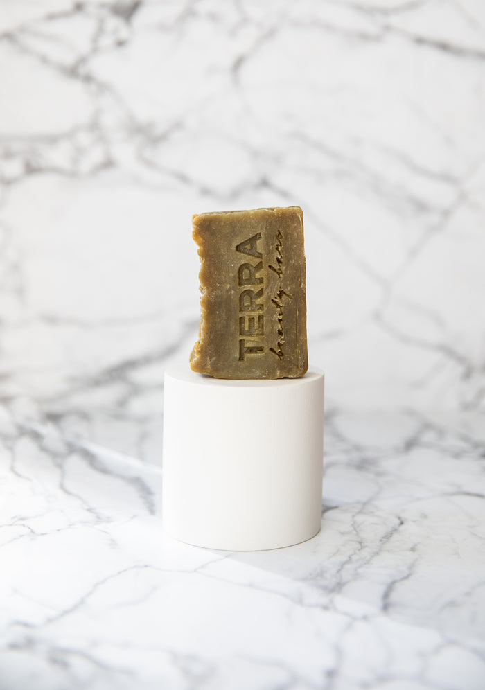The Clean Green Tea Tree Facial Beauty Bar, engraved with TERRA and infused with rejuvenating Green Sea clay, stands vertically on a white cylindrical pedestal against a white marble background.