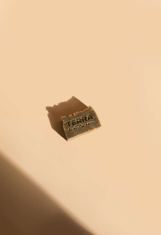 A small, rectangular Clean Green Tea Tree Facial Beauty Bar from Terra Beauty Bars lies on a beige background with a delicate dusting akin to matcha tea, casting a subtle shadow.